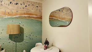 Ein Zimmer mit Wandgemälde zum Strandthema, einem runden Spiegel, einem kleinen Tisch mit einem Getränk und einer Lampe.