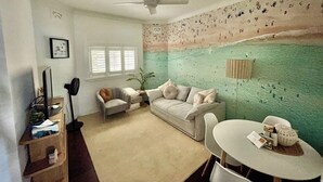 Ein Zimmer mit Deckenventilator, beiges Sofa, kleinem Tisch und Wand mit Strandbild.