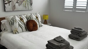Ein ordentlich bezogenes Bett mit einer weißen Daunendecke, zwei Kissen mit Vogelmotiven und einem braunen Zierkissen, sowie ein Stapel gefalteter Handtücher.