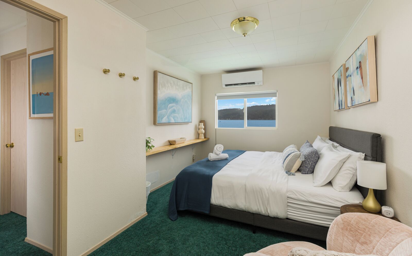 A bedroom with a bed, a nightstand, a lamp, a window with a view, an air conditioning unit, and a door.