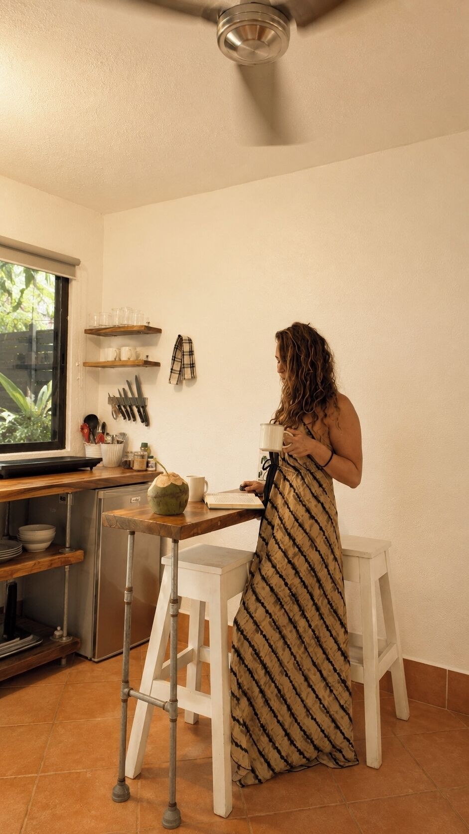 A woman in a striped dress at a kitchen counter with a laptop, a mug, and a green pot. There are white stools, a window with a view, and kitchen utensils on the wall.