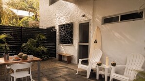 Une terrasse couverte avec une table et des chaises, une planche de surf et une fenêtre ornée d’une grille métallique.