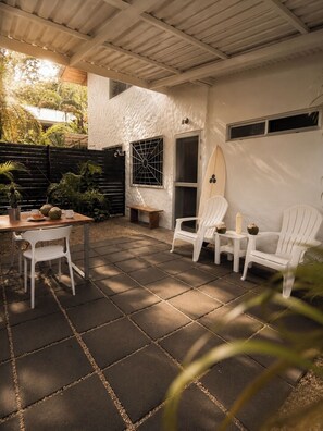 A covered patio with a table and chairs, a surfboard, and a window with a metal grid. - SALT 2 - 1BR with private patio in Guiones Nosara (Playa Guiones)