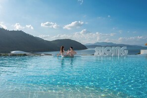 Una pareja sentada en una piscina infinita de color azul intenso, con vistas a una montaña y al mar.