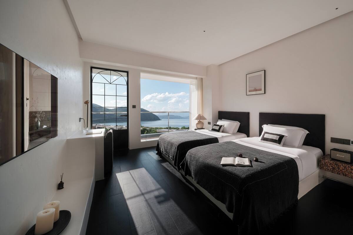 Ein modernes Schlafzimmer mit zwei Betten, einem großen Fenster mit Meerblick und minimalistischem Design.