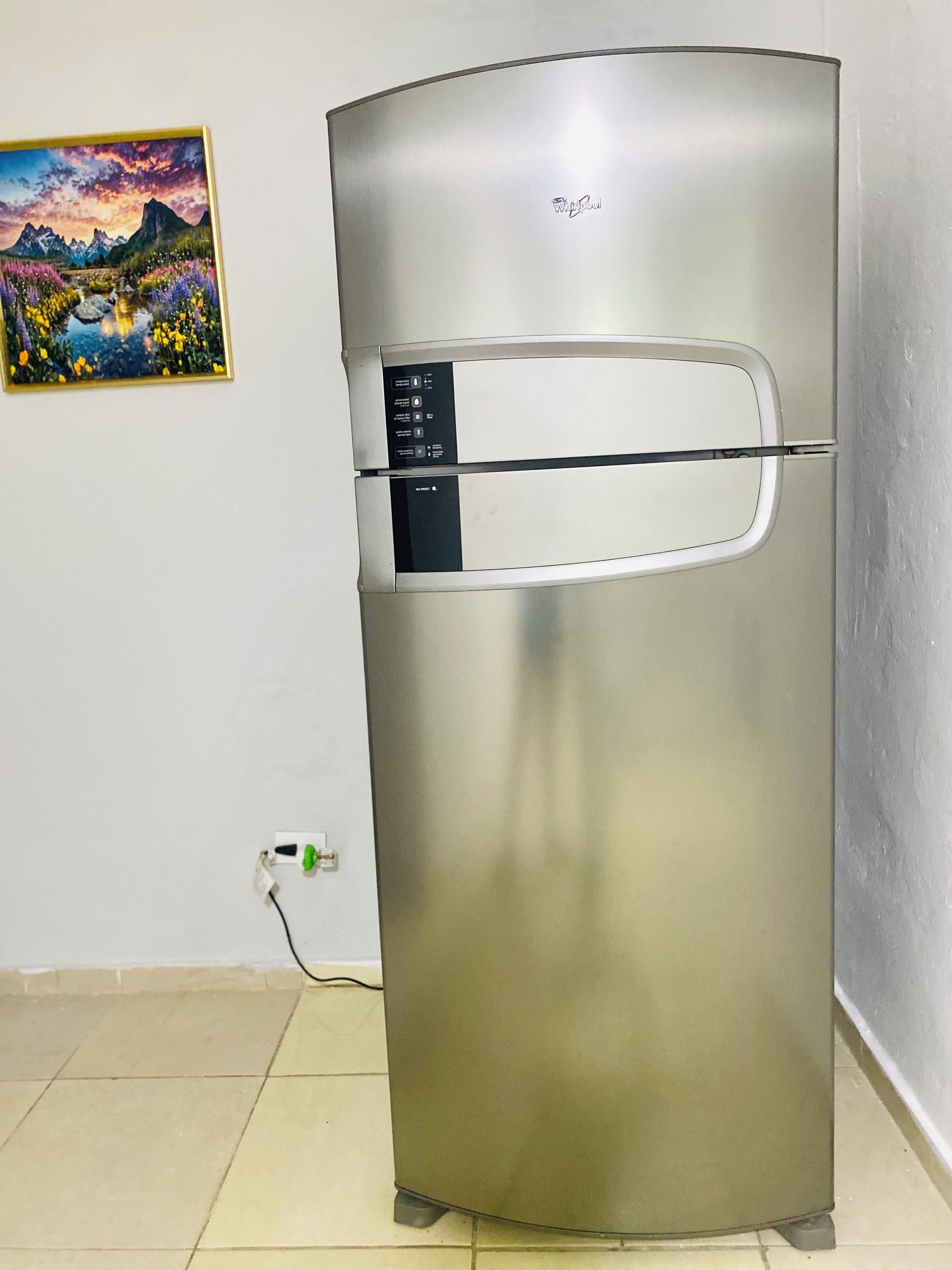 A stainless steel refrigerator with a control panel, a power cord, and a wall-mounted picture.