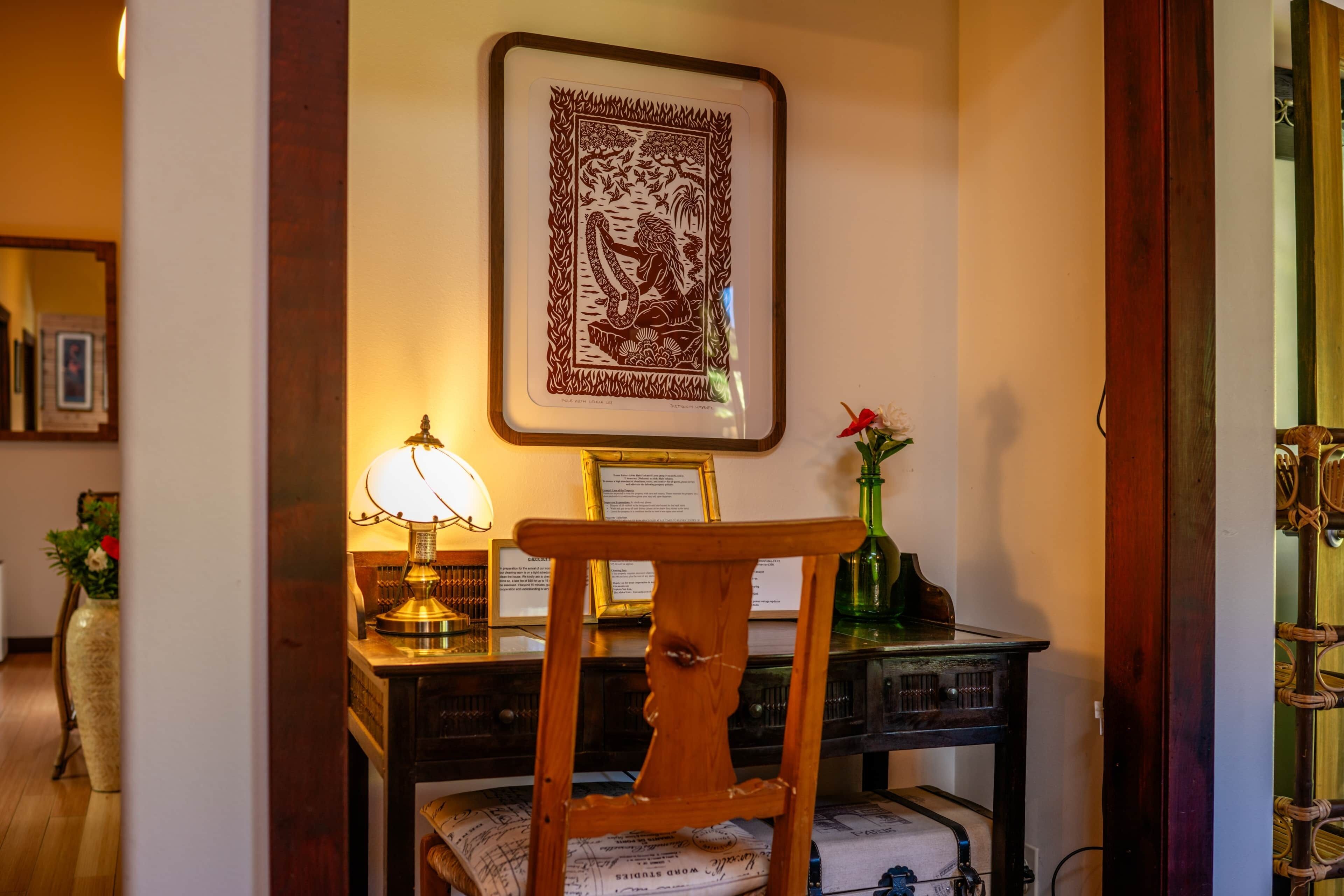 A room with a wooden chair, a desk with a lamp, a framed artwork, and a vase with flowers.