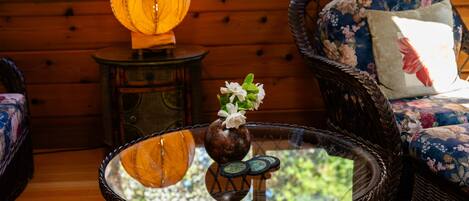 Uma mesa de centro de vime com tampo de vidro, um vaso com flores brancas e um candeeiro num apoio lateral de madeira.