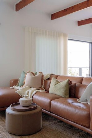 A living room with a brown leather sofa, a round ottoman, and a window with sheer curtains.