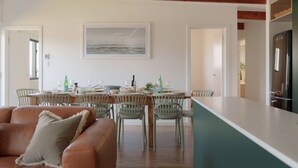 A dining area with a wooden table and chairs, a kitchen with a refrigerator, and a living room with a leather sofa. - Soul Shack ~ 6 bedroom Soulful Coastal Escape (Aldinga Beach)