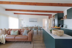 A modern living room with a leather sofa, dining area, kitchen island, and wooden beams on the ceiling. - Soul Shack | Pet Friendly + Steps to Beach (Aldinga Beach)