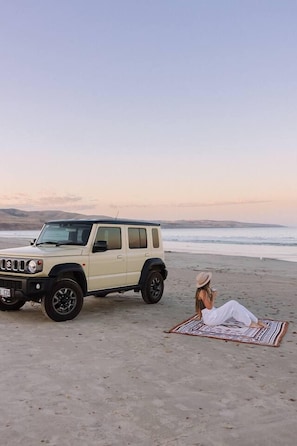 A beige SUV parked on a beach with a person lying on a patterned blanket. - Soul Shack | Pet Friendly + Steps to Beach (Aldinga Beach)