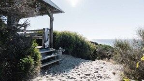 A wooden beach hut with a metal roof, surrounded by sand and sparse vegetation, overlooking a clear sky and calm sea. - Kestrels Nest - a luxurious couples retreat (Aldinga Beach)