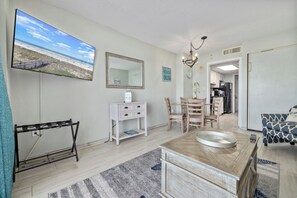 A modern, well-lit room with a large TV displaying a beach scene, a wooden vanity with a sink, a dining area with chairs, and a kitchen visible through an open door. - Baywatch 1734: Family fun pools & lazy rivers (North Myrtle Beach)