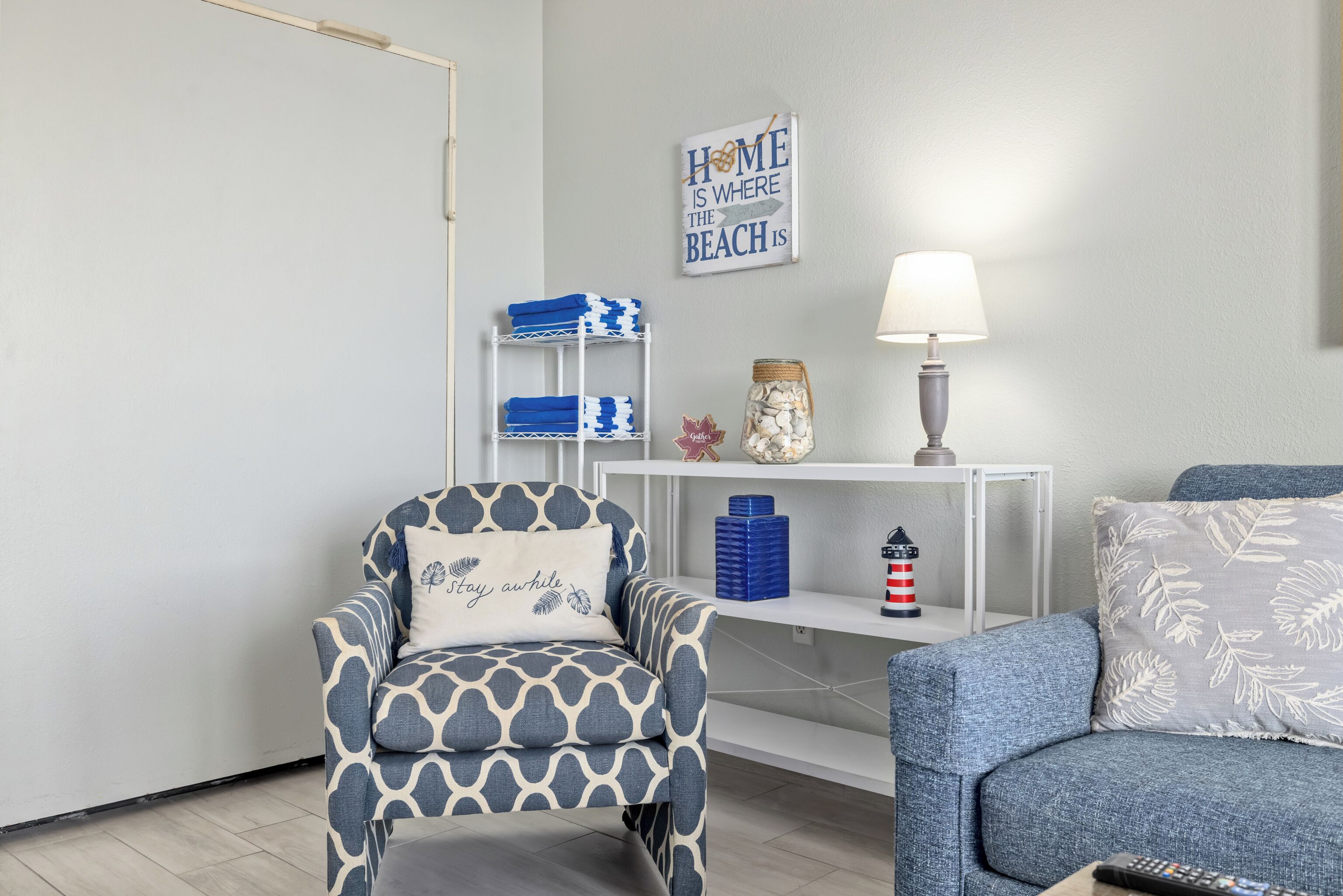 Un salón con un sillón estampado, un sofá azul, una consola blanca con adornos, y un letrero en la pared.