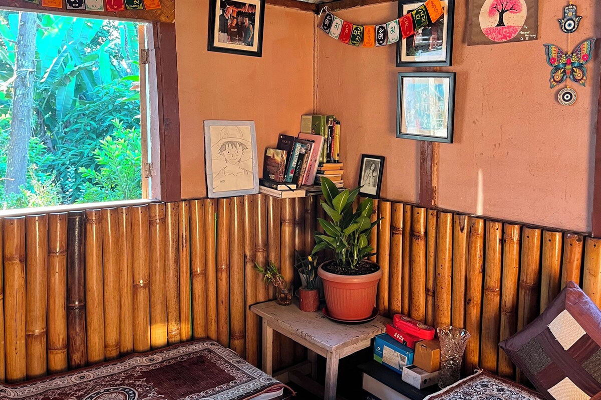 A room with a bamboo wall, a bed with a colorful quilt, a small table with a potted plant, and a window with a view of greenery.