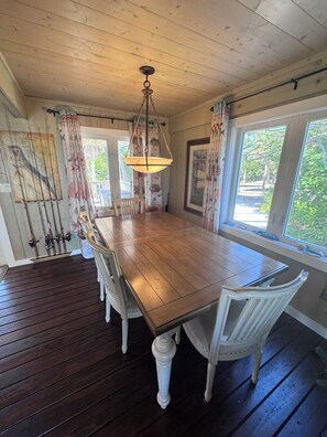 Uma área de refeições com uma mesa de madeira, cadeiras brancas, um lustre e janelas com cortinas.