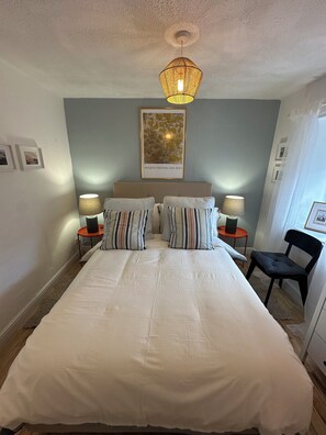 Un dormitorio con una cama grande, dos mesitas de noche con lámparas, una silla, un candelabro en el techo y un cuadro enmarcado en la pared.