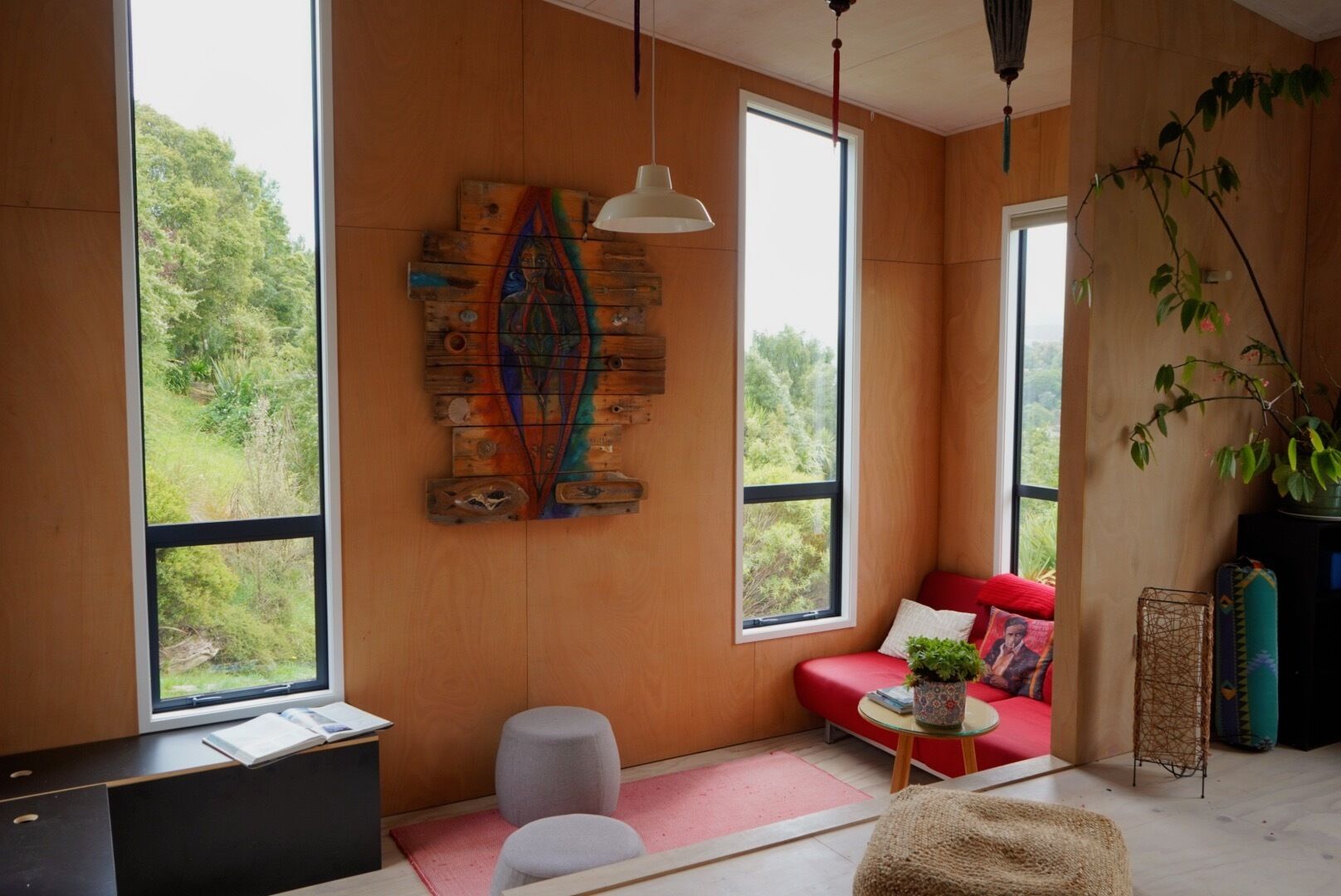 A cozy room with large windows, a red sofa, a small table with a plant, and a unique wooden artwork on the wall. - Funky Eco Pad - Innovative Design, with great indoor/outdoor flow.  Pet Friendly (Nelson)