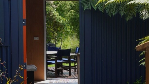 A doorway leading to an outdoor seating area with a table and chairs, surrounded by greenery and a corrugated metal wall. - Funky Eco Pad - Innovative Design, with great indoor/outdoor flow.  Pet Friendly (Nelson)