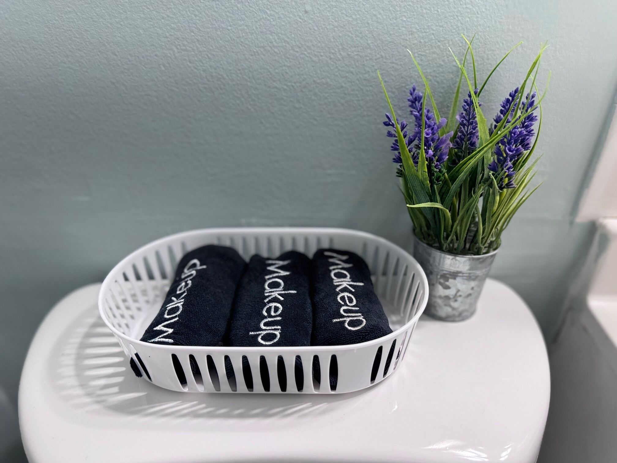 A white toilet with a white basket containing three dark blue towels with the word "makeup" on them. There is a small potted plant with purple flowers to the right.