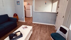 A living room with a white coffee table, a blue sofa, and a white chair. There is a kitchen visible through an open door. - Surf Shack Across from the Beach (Cocoa Beach)
