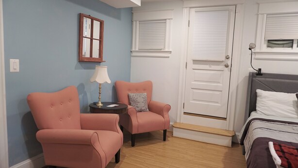 A bedroom with two pink armchairs, a bedside table with a lamp, a bed with a white headboard, and a door with a window above it. - Charming & Convenient Suite in Petworth w/ Parking (Washington)