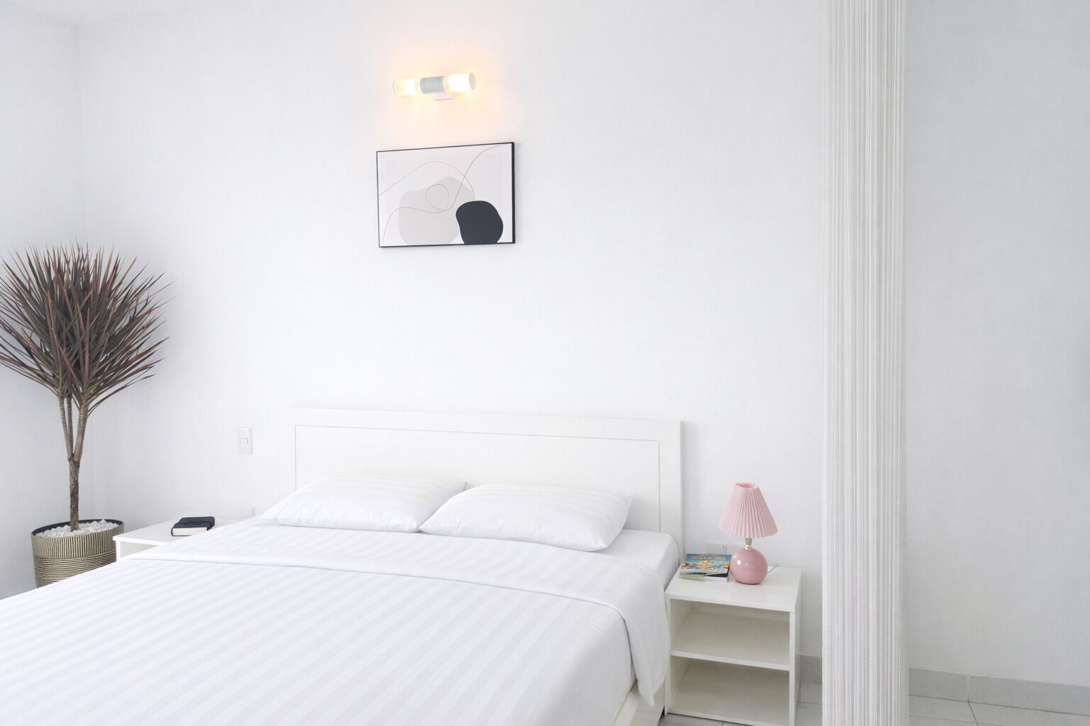 Une chambre à coucher au style minimaliste, avec un lit blanc, une table de chevet dotée d’une lampe et un luminaire fixé au mur.