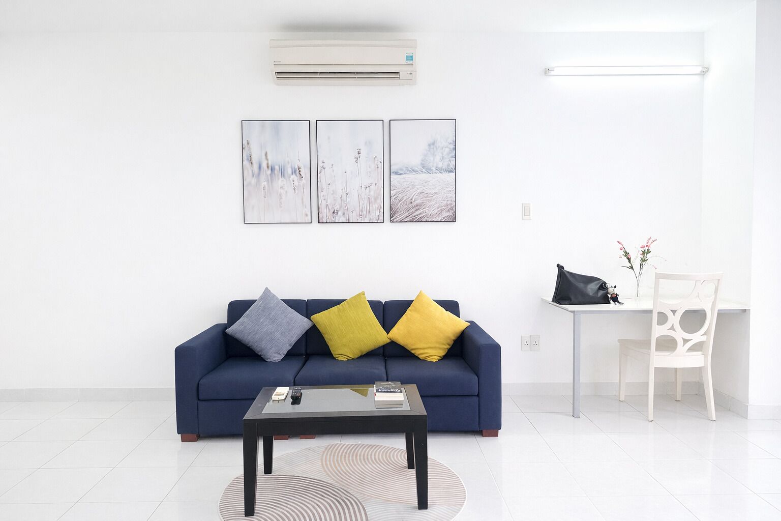 A modern living room with a blue sofa, a coffee table, and a white chair. There are three framed pictures on the wall and an air conditioning unit above.