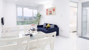 A modern living room with a glass dining table, white chairs, a dark blue sofa, and a large window with a view of greenery. - HD Garden Thu Dau Mot (Ho Chi Minh City)