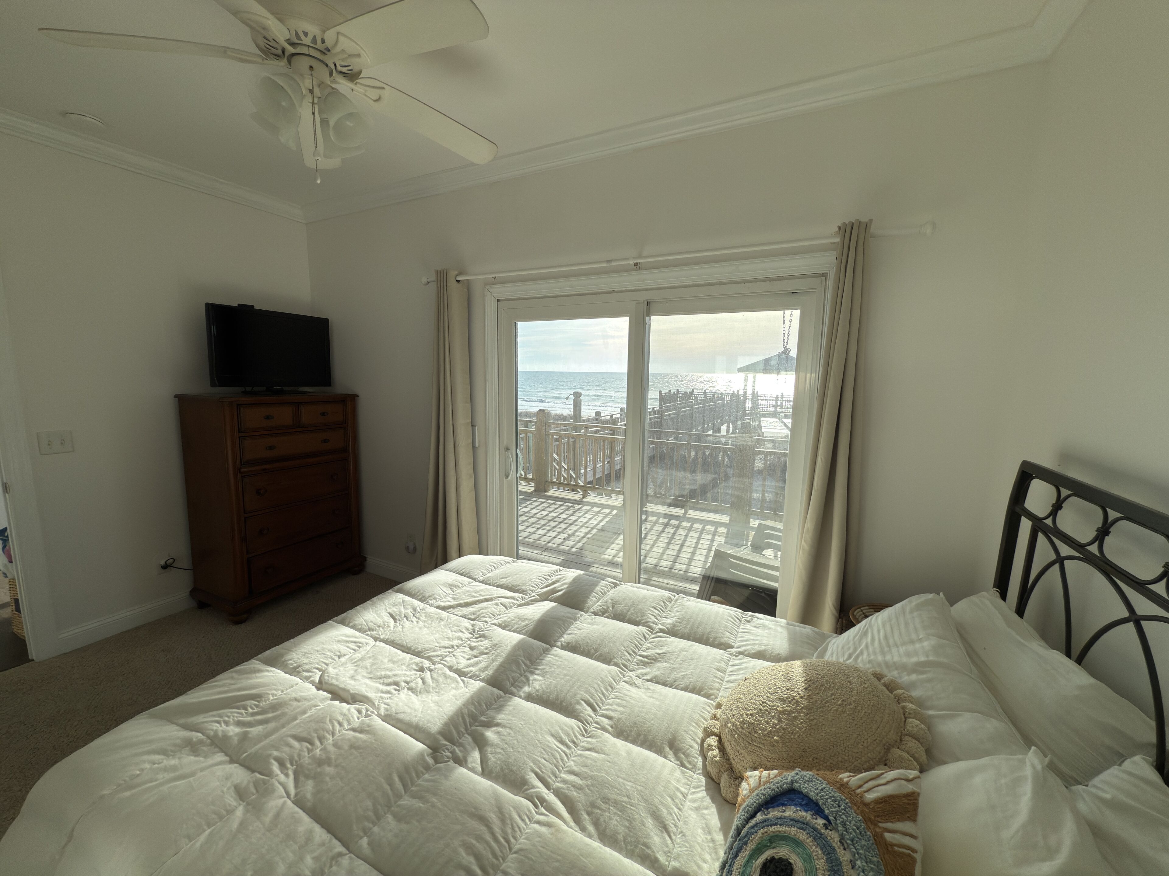 A bedroom with a bed, a dresser, a television, and a balcony with a view of the city and the sea.
