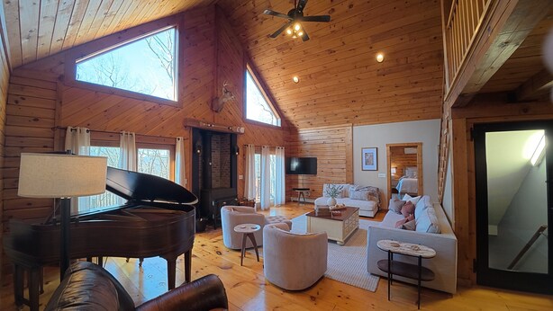 A cozy cabin interior with a grand piano, comfortable seating, a fireplace, and large windows.