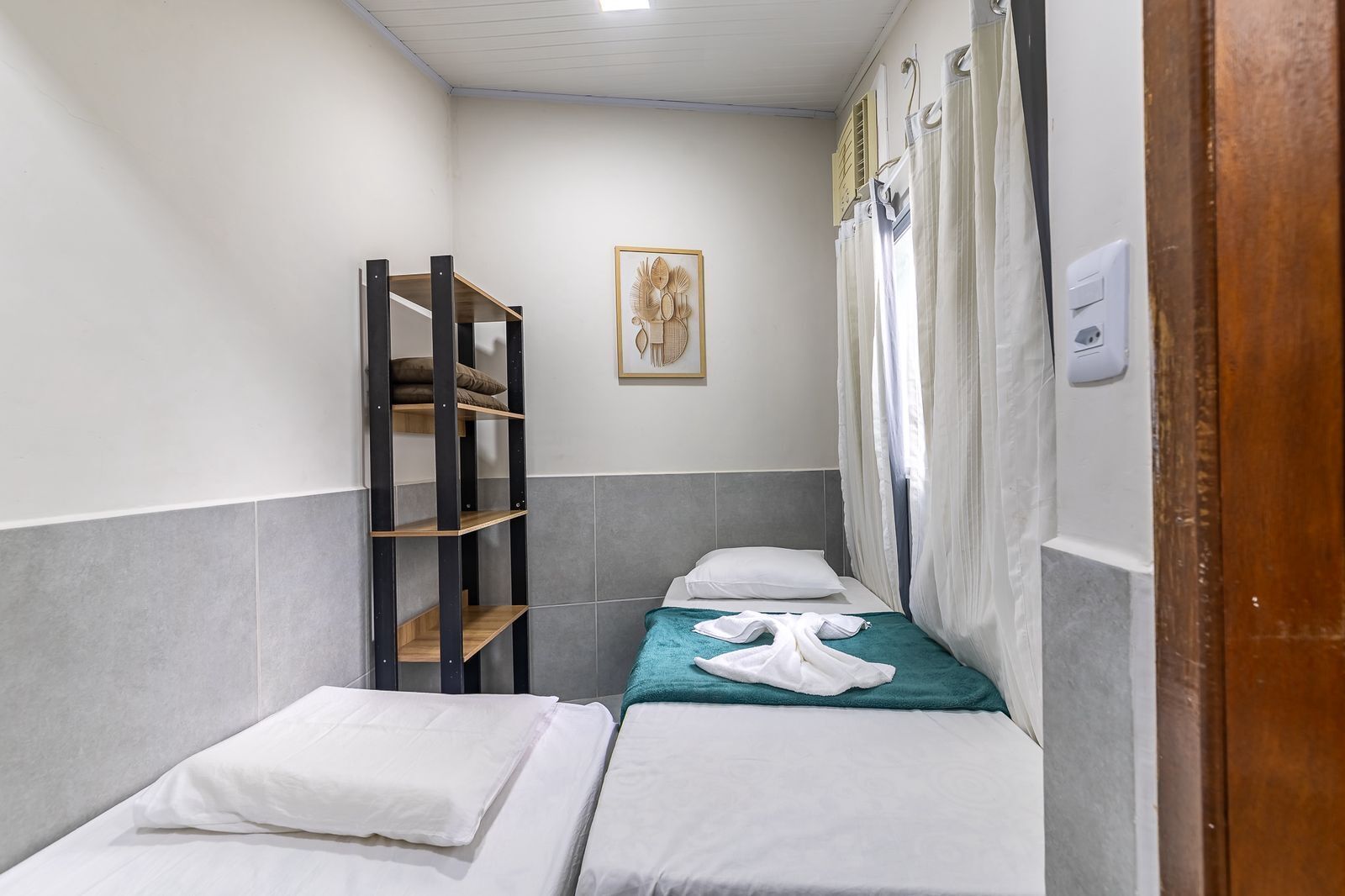 Une petite chambre d’hôtel étroite, avec deux lits simples, une étagère en bois, une œuvre d’art encadrée au mur et une fenêtre avec des rideaux.