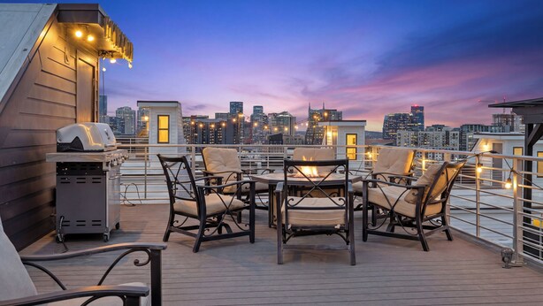 A rooftop terrace with outdoor seating, a grill, and a city skyline view at dusk.