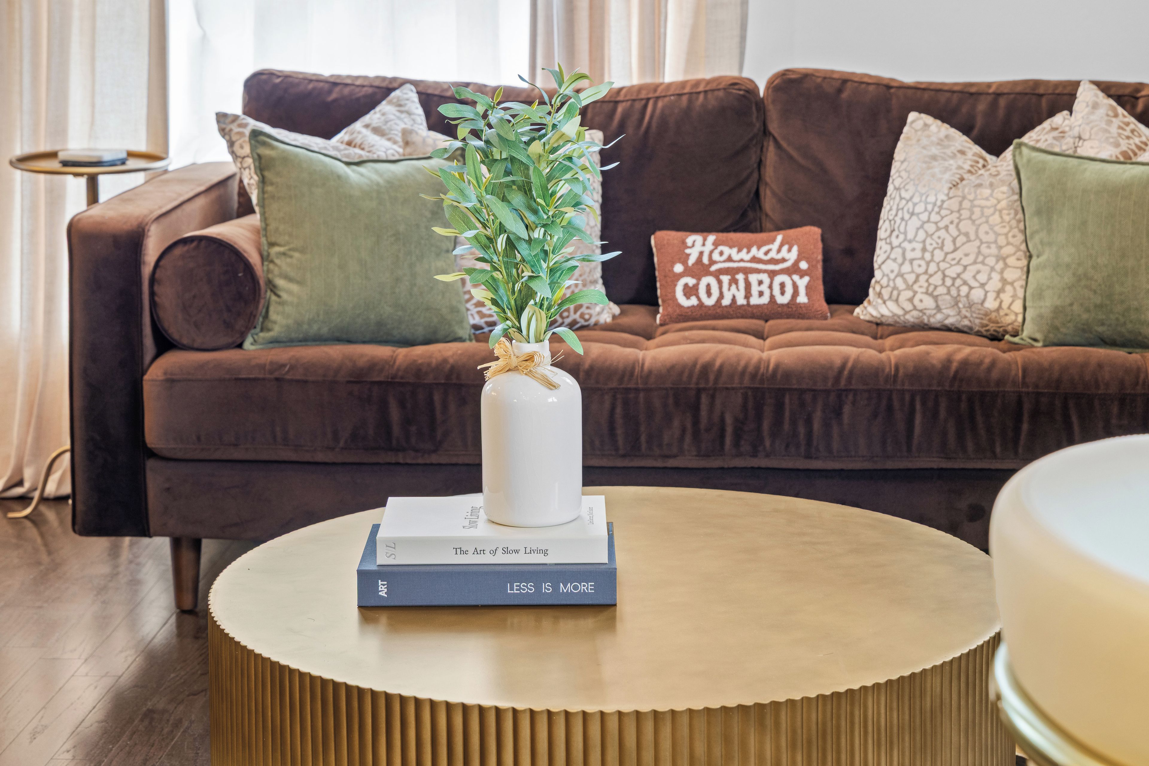 A brown sofa with a decorative pillow, a white vase with green foliage, and a book titled 'The Art of Slow Living' on a round wooden table.