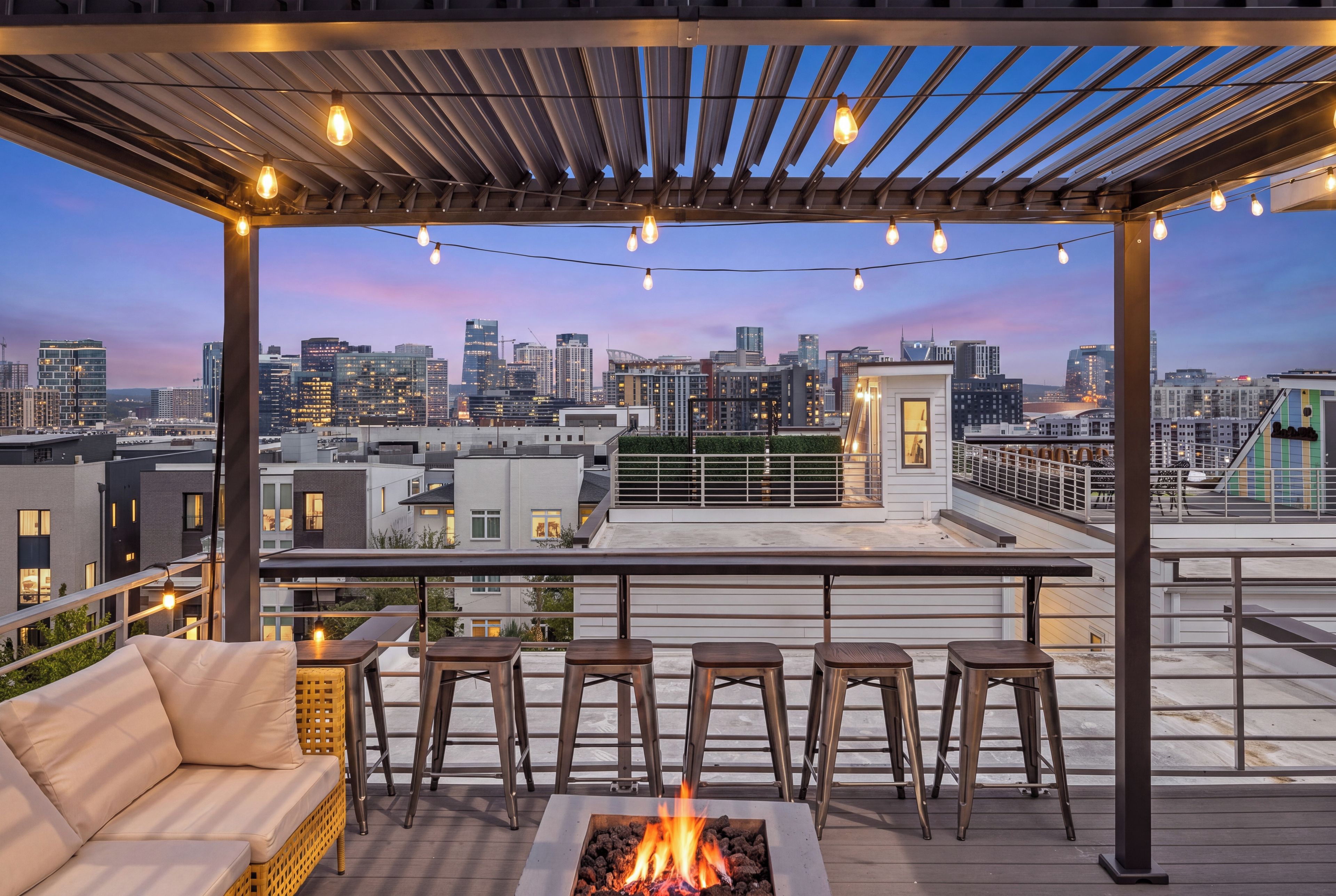 A rooftop terrace with a fire pit, seating, and a city view at dusk.
