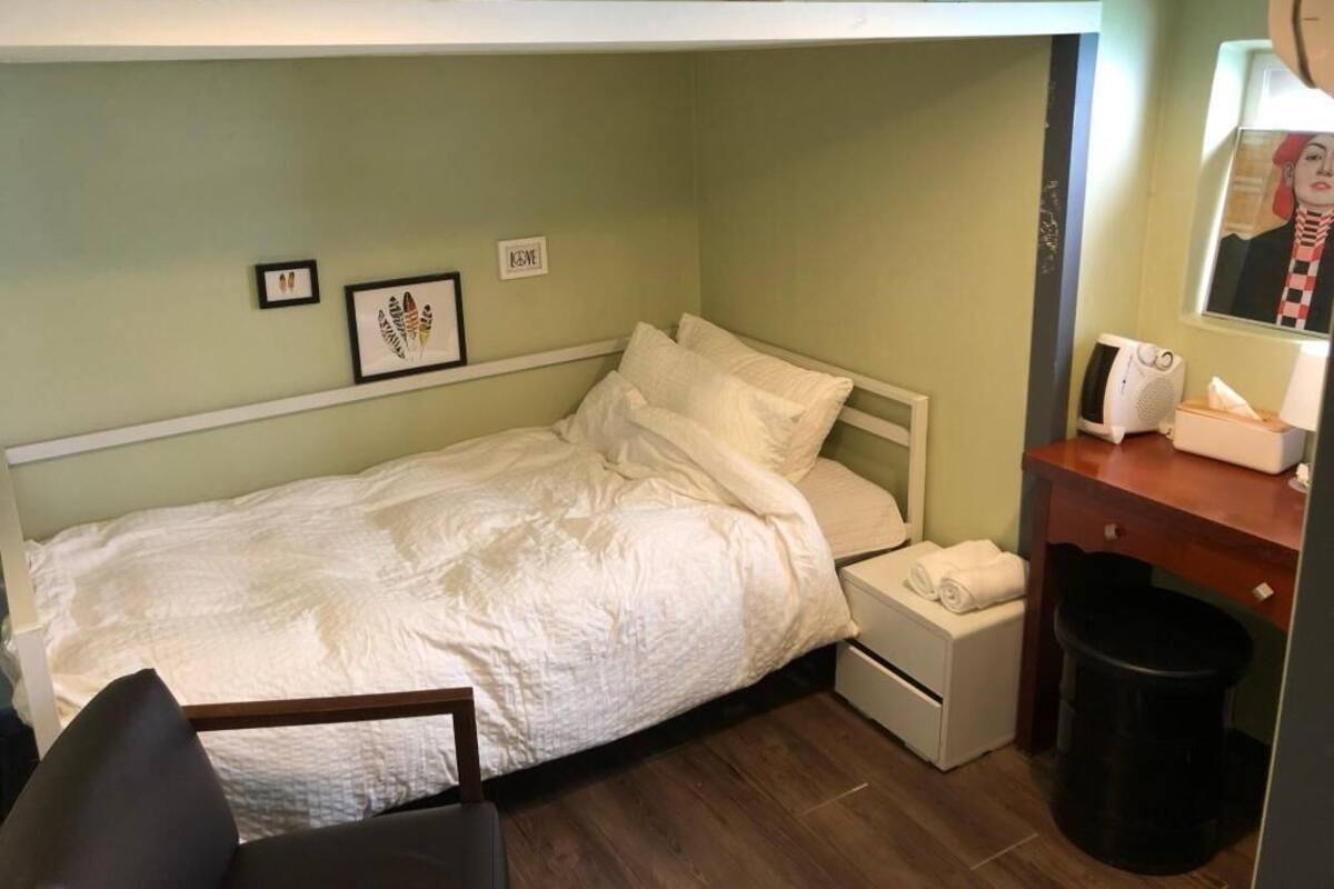 A small, single-bed room with a white bed, a nightstand, a chair, a small desk with a mirror, and a shelf with framed pictures.