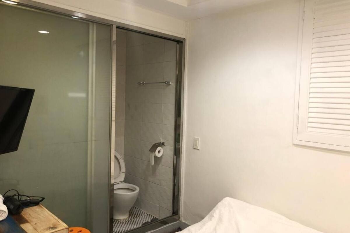 A bathroom with a glass shower enclosure, a toilet, and a white bed.