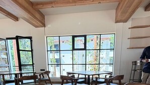 A dining area with wooden tables and chairs, large windows with a view of buildings and trees, and a ceiling fan. - Samarya CHikkamagaluru (Chikkamagaluru)