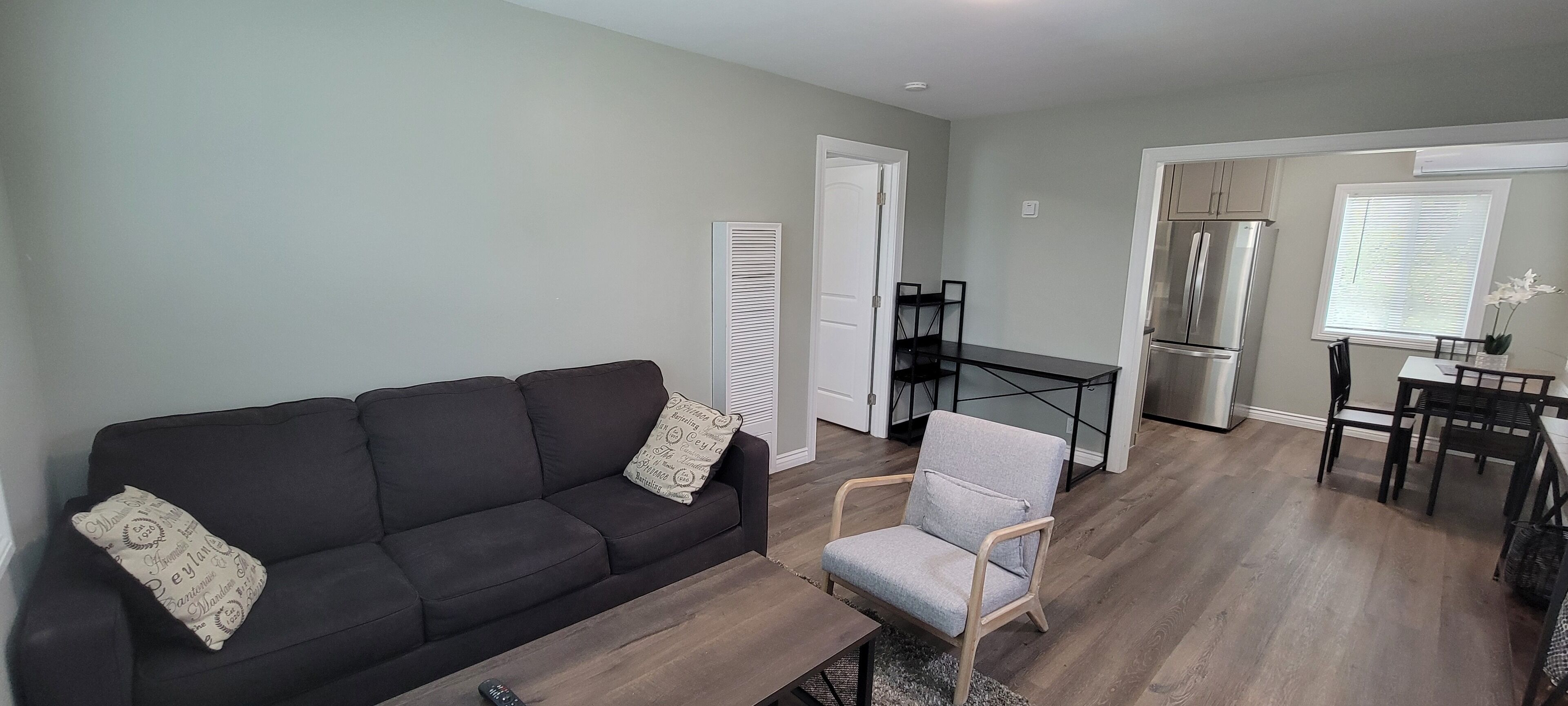 Ein Wohnzimmer mit einem schwarzen Sofa, einem grauen Sessel, einem Holztisch und einem Essbereich mit Tisch und Stühlen.