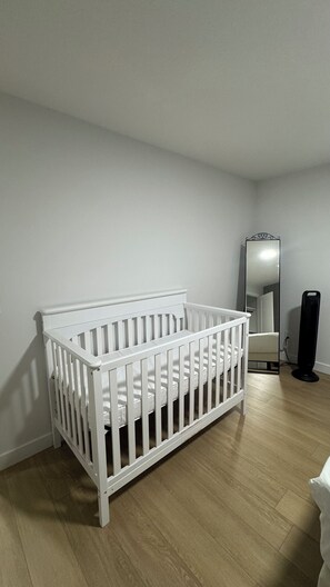 A white crib in a room with a wooden floor, a mirror, and a black fan.