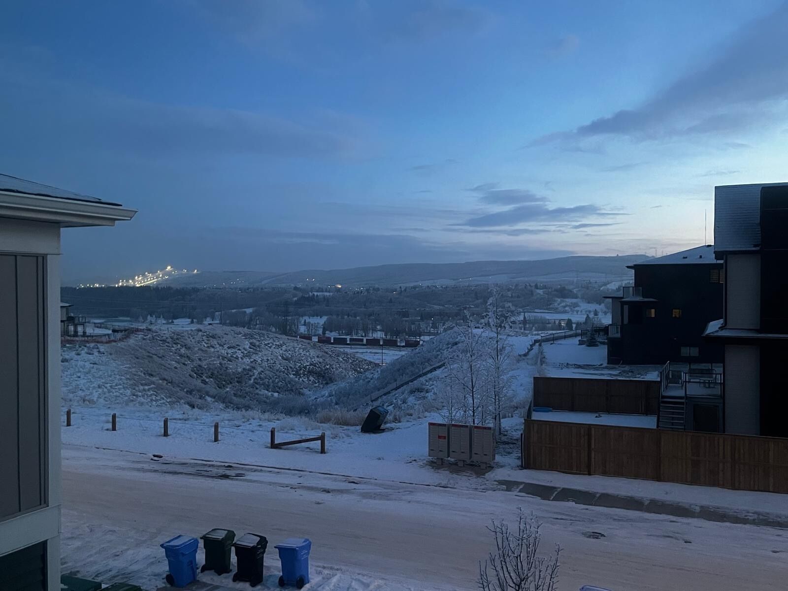 Zona residencial con suelo cubierto de nieve, vistas a luces distantes y un edificio moderno con techo plano.