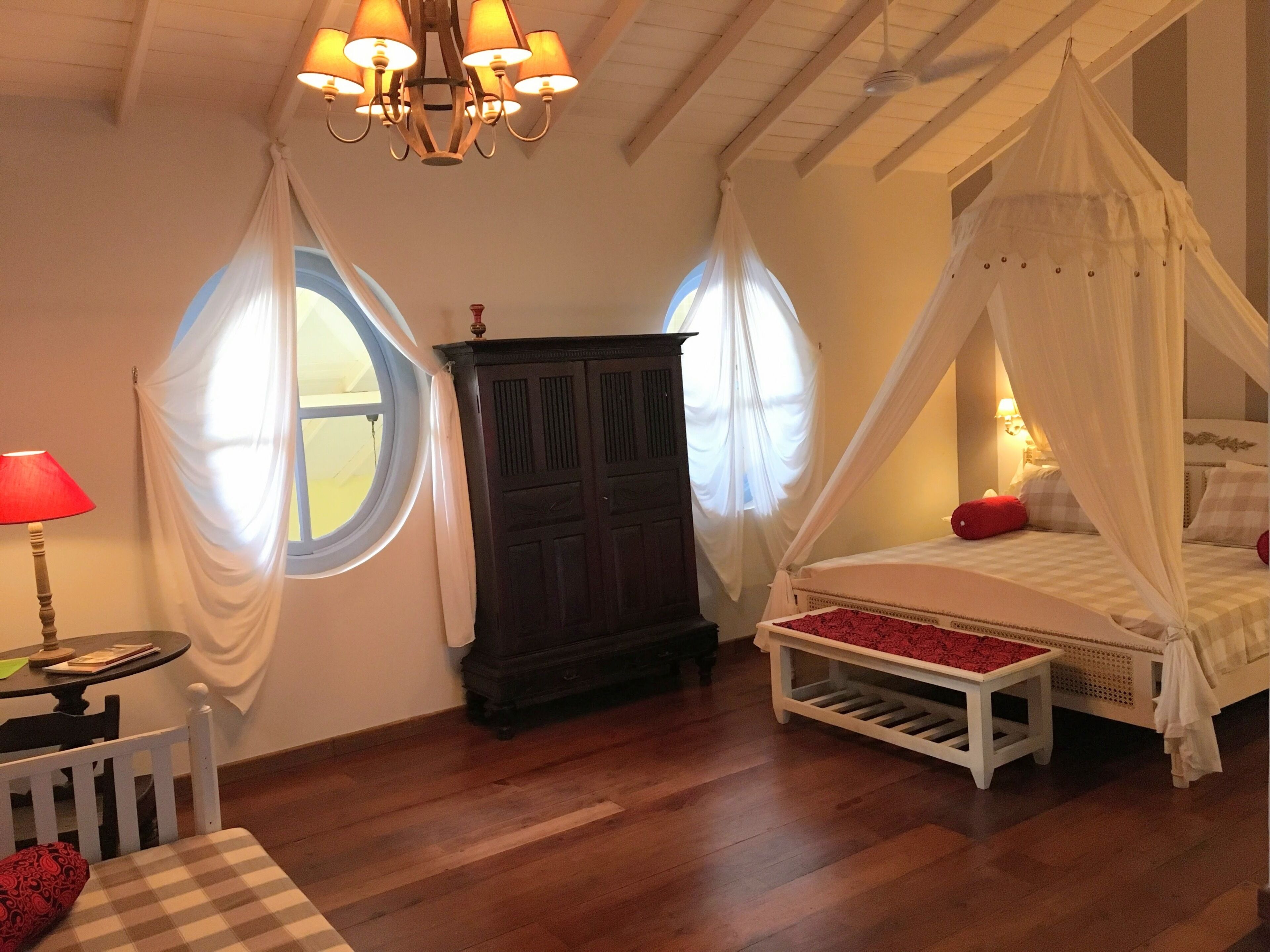 Quarto com cama de dossel, guarda-roupa de madeira, mesa redonda com abajur e duas janelas com cortinas.