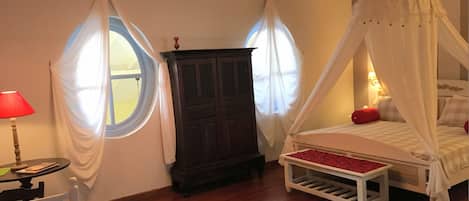 Quarto com cama de dossel, guarda-roupa de madeira, mesa redonda com abajur e duas janelas com cortinas.