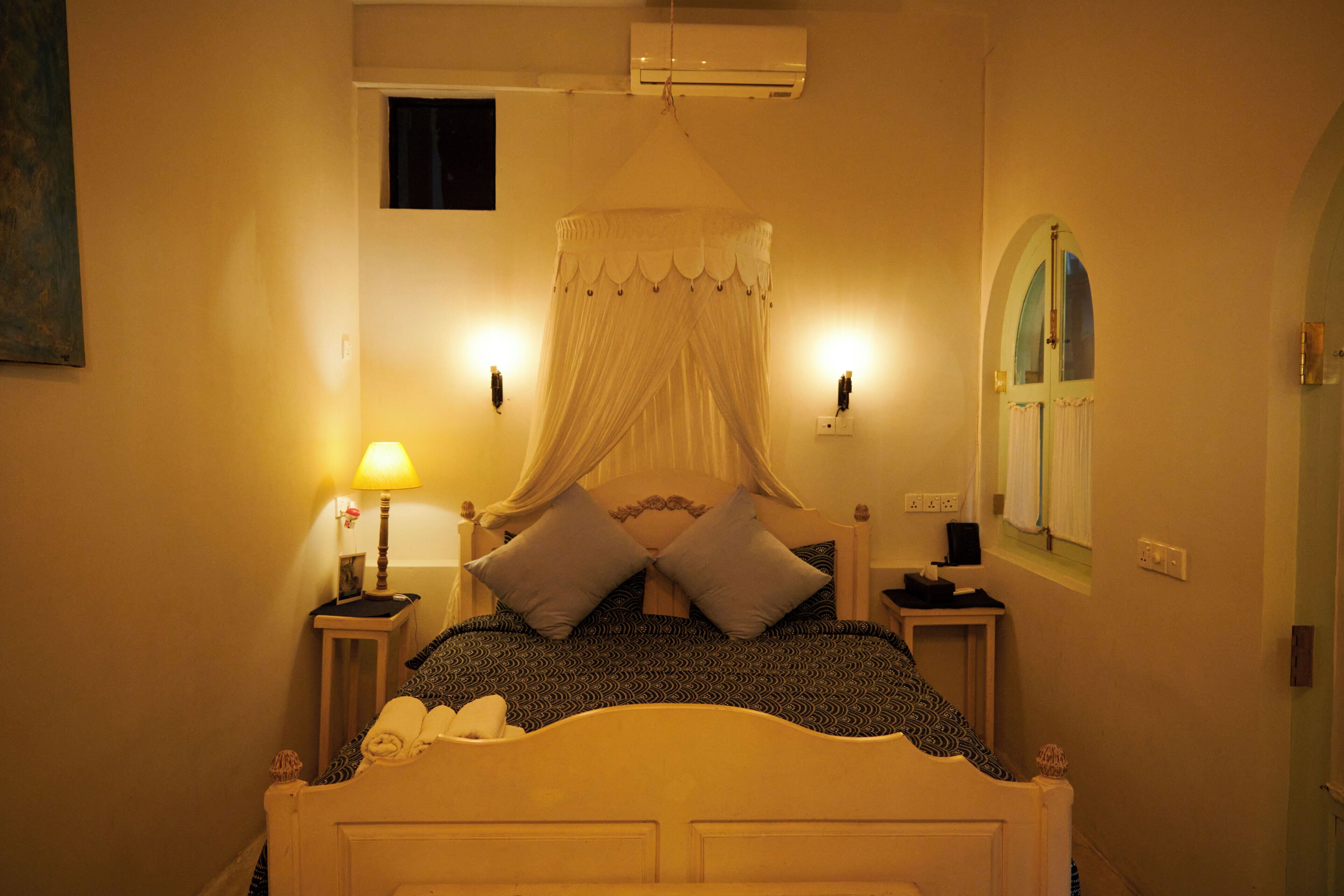 A bed with a canopy, bedside lamps, a wall-mounted air conditioner, and a window with a curtain.