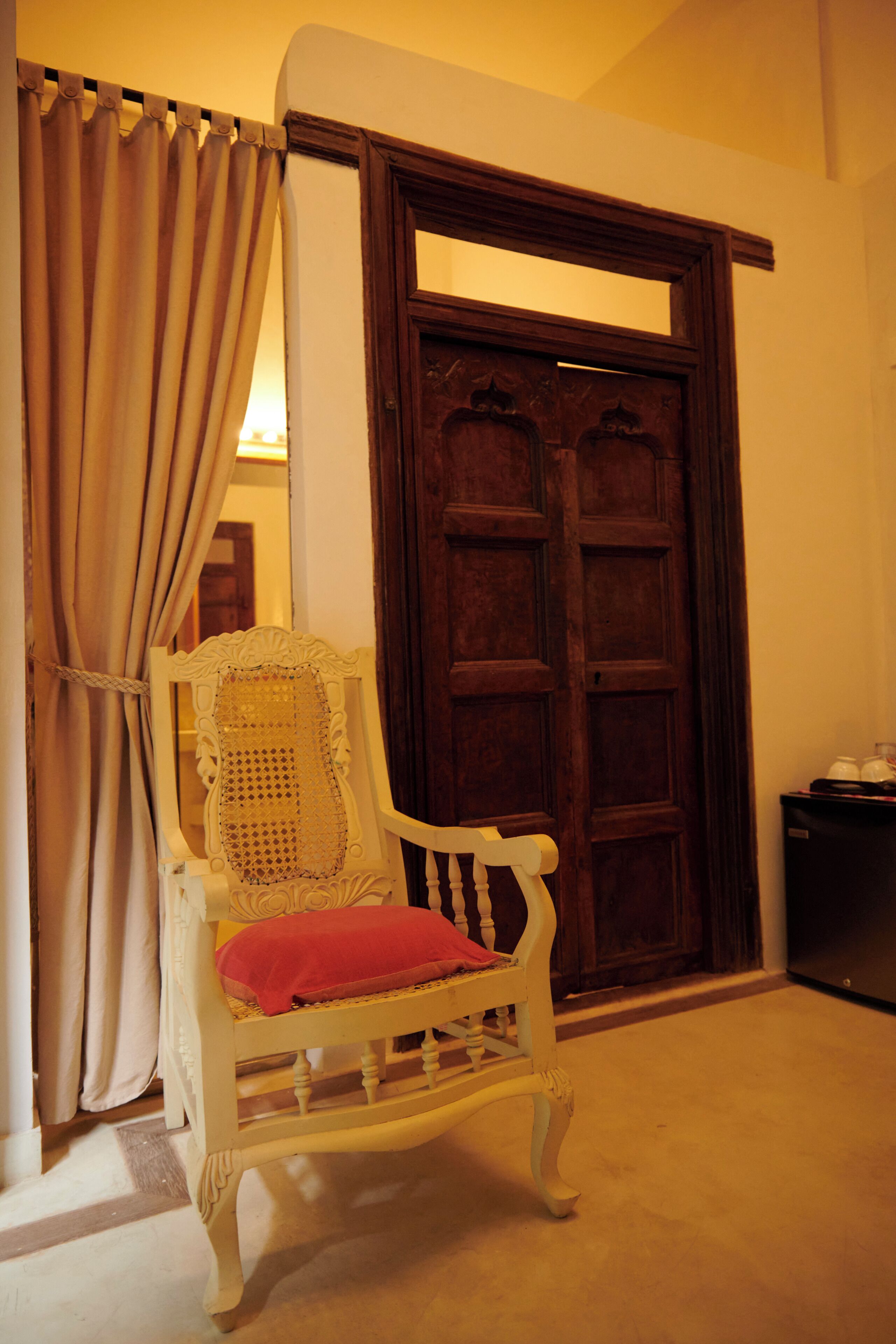 A room with a wooden door, a white chair with a red cushion, and a curtain.
