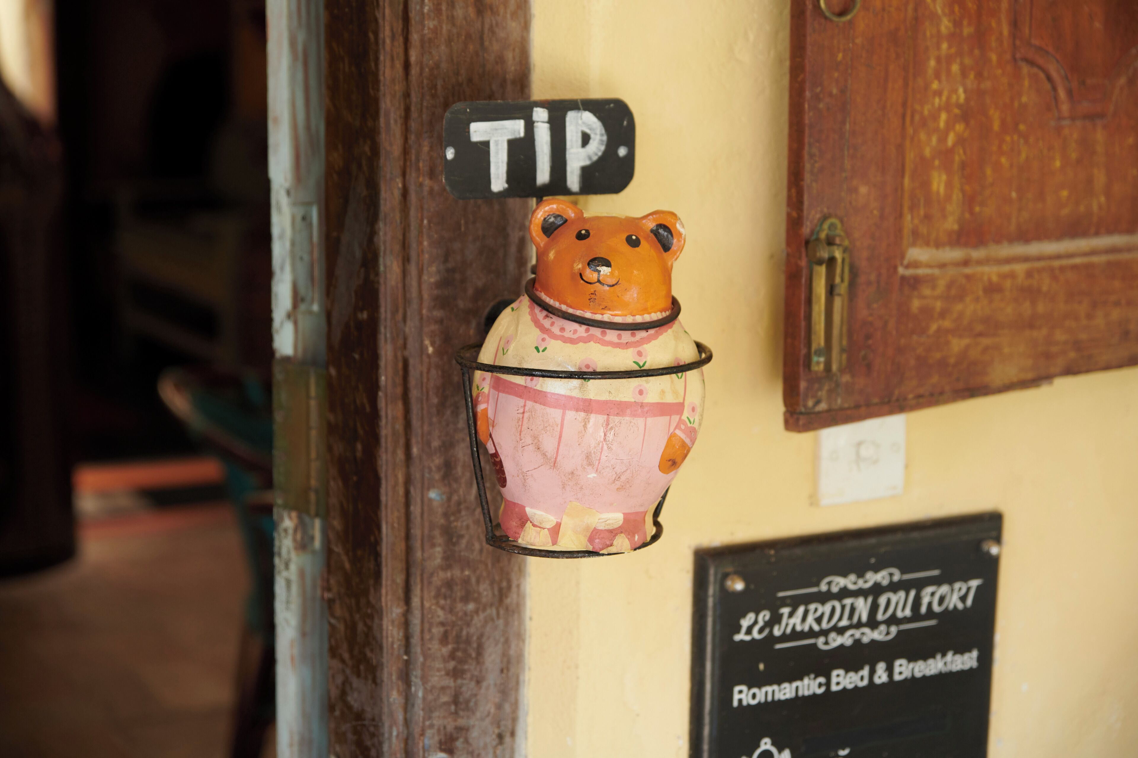 A bear-shaped tip jar on a door, a sign for 'Le Jardin du Fort' offering romantic bed and breakfast, and a wooden door with a brass handle.