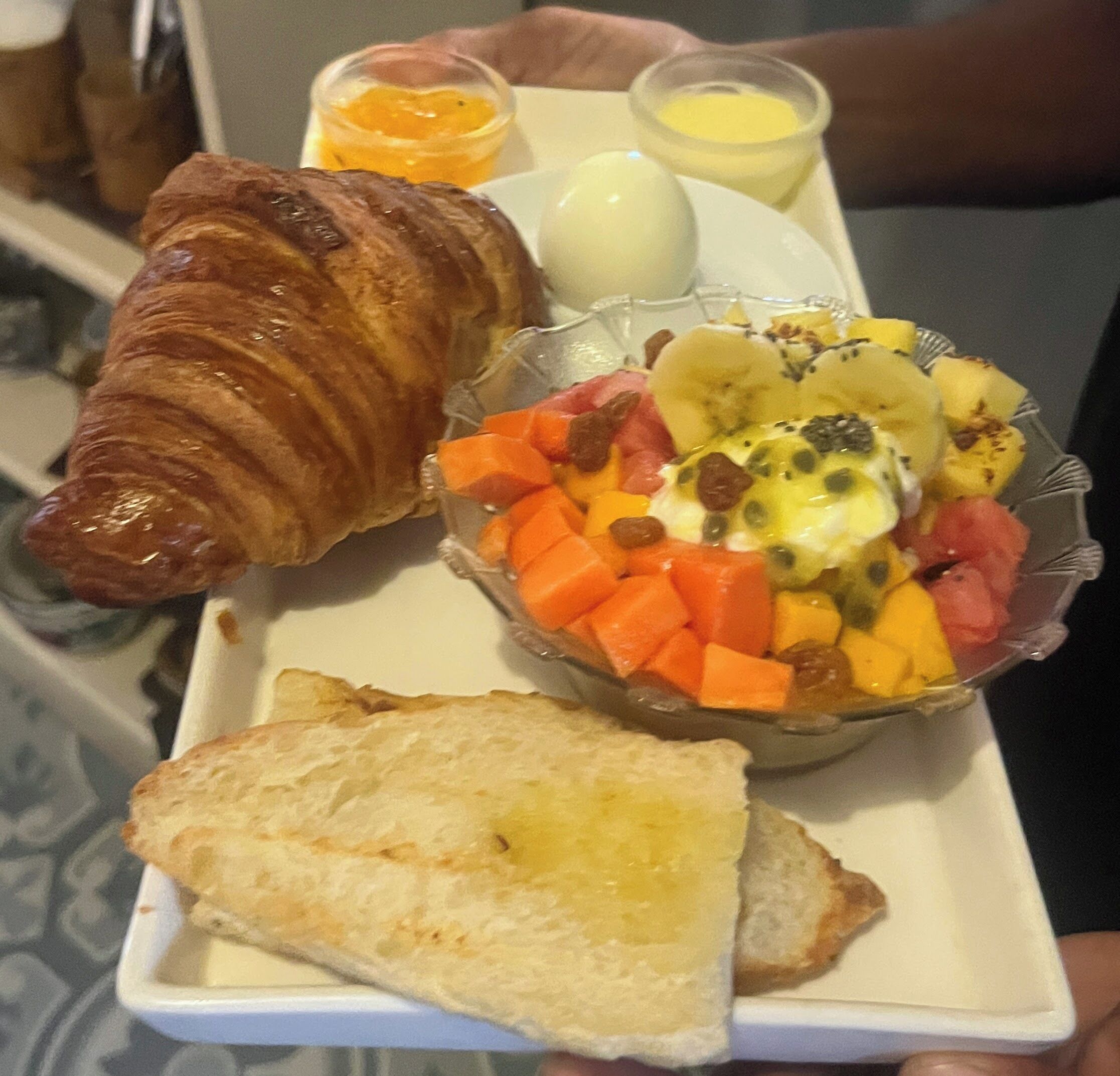 Una bandeja con un croissant, una ensalada de frutas, un huevo duro y dos tostadas.