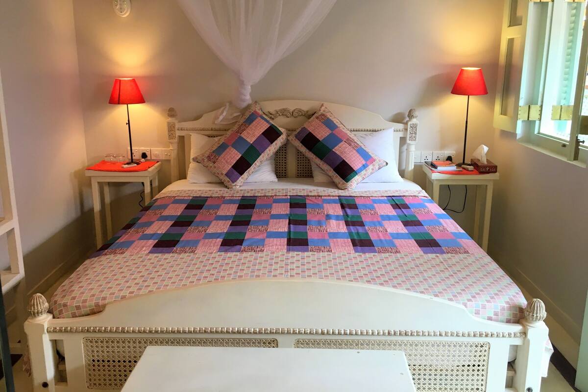 Un lit bien fait, recouvert d’une couvre-lit à carreaux, deux lampes de chevet, une table de chevet avec une boîte de mouchoirs et une fenêtre avec des rideaux.