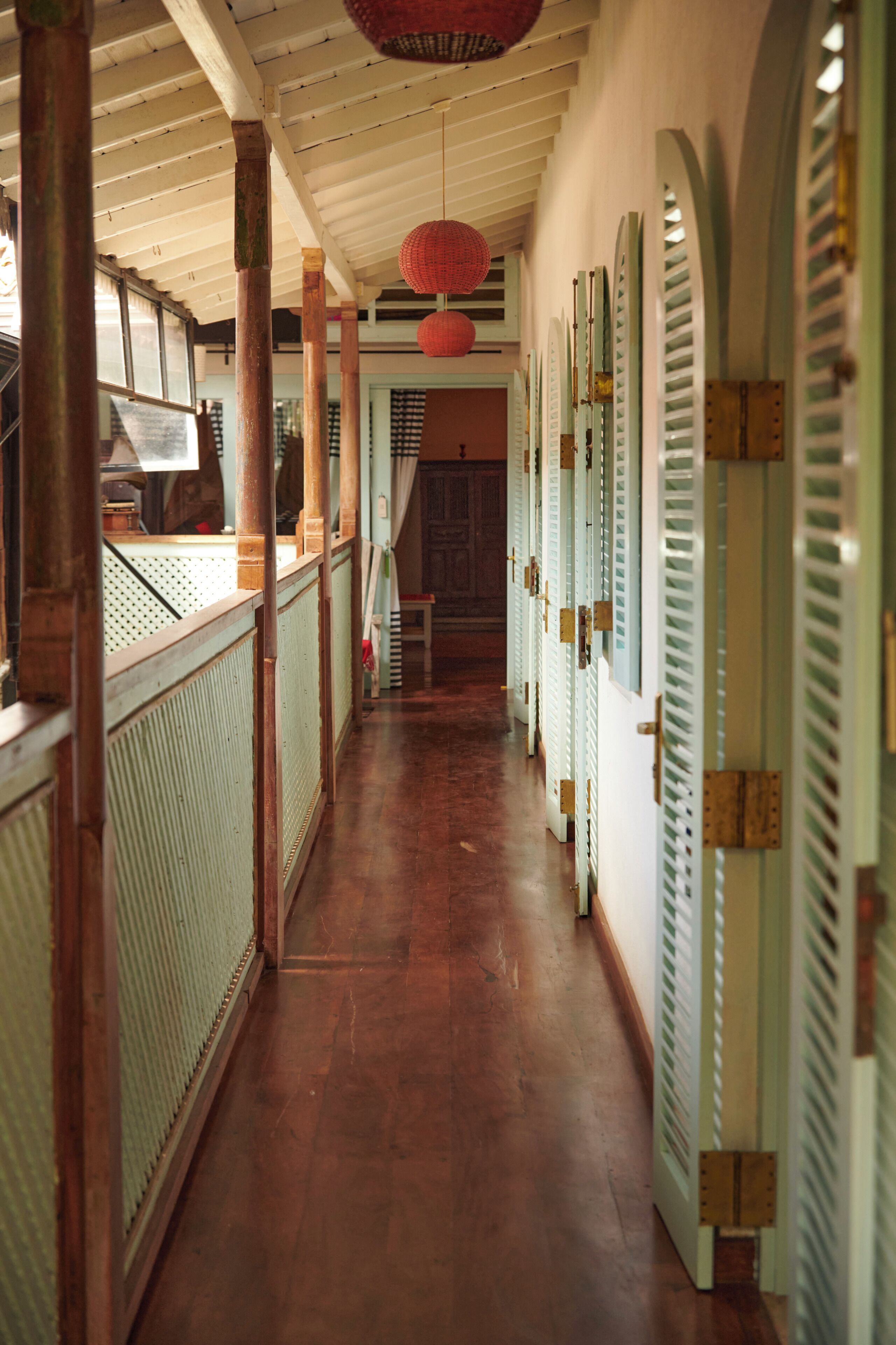 A hallway with wooden flooring, a ceiling with a hanging red lantern, and a row of doors with green shutters.